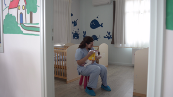 A nurse sits on a stool in the middle of a baby room and feeds a baby in her arms